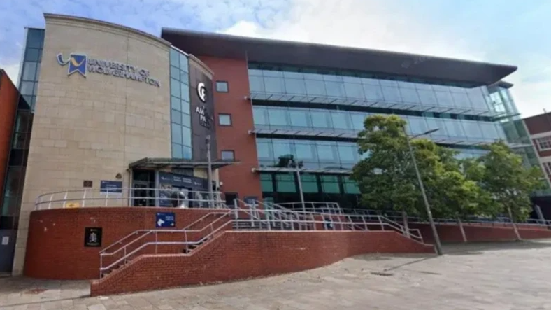 The front of the university is glass, red brick and stone. There is a railed walkway up to the entrance and some trees on the right hand side.