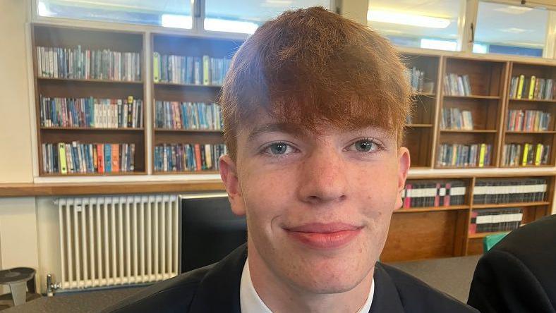 Corey has red hair with a long fringe brushed forward and is wearing a black blazer and white shirt