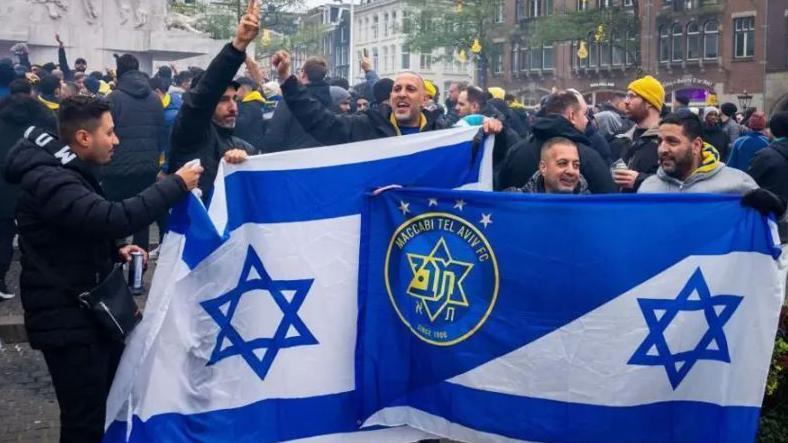 Fans of Maccabi-Tel Aviv hold club flags as they sing out in support of their club, as other fans are gathered behind them, in an Amsterdam square.