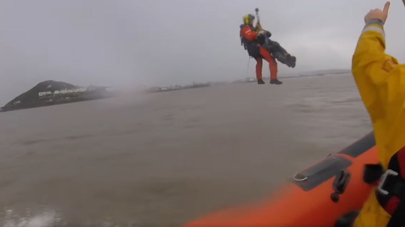 An RNLI crew member being winched above the water with the casualty strapped to him and the edge of a lifeboat in the foreground