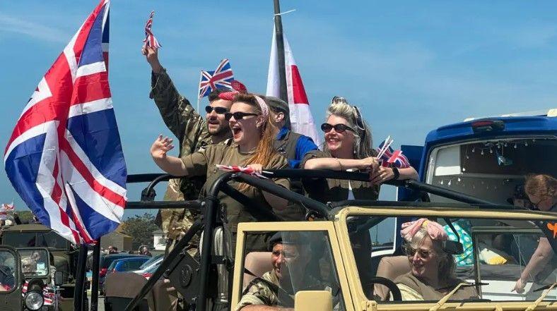 Close up of cavalcade. Women and men are in the back of a car and are dressed up in khaki military green outfits. They are waving Union Jack flags.  