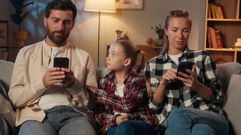 Girl sat on sofa between her parents who are both on their phones, trying to get the attention of her father.