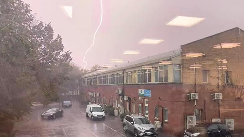 A lighting bolt cuts through the sky down the middle of this image. A red brick building with cars alongside can also be seen. The sky is grey in what appears to be a torrential downpour.