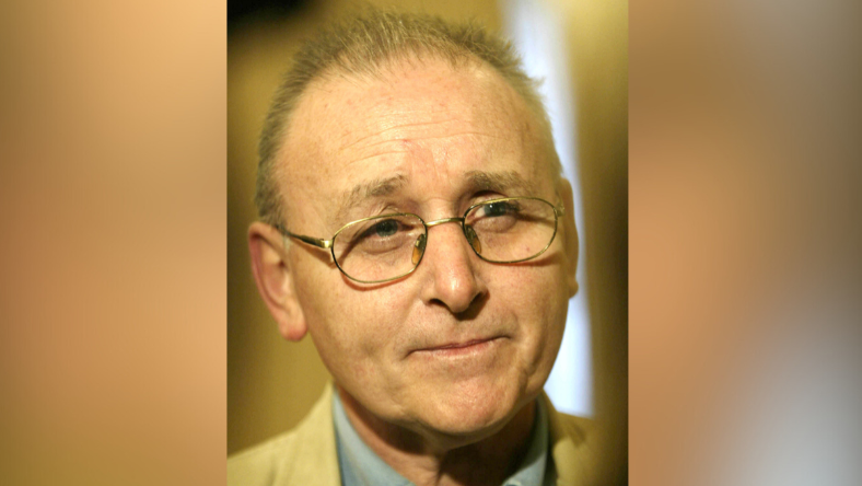 A close up image of Denis Donaldson. He has thinning, short grey hair and has a neutral expression on his face as he takes a question at a press conference. He has a pair of silver glasses on. He is wearing a tan suit jacket and a blue collared shirt.