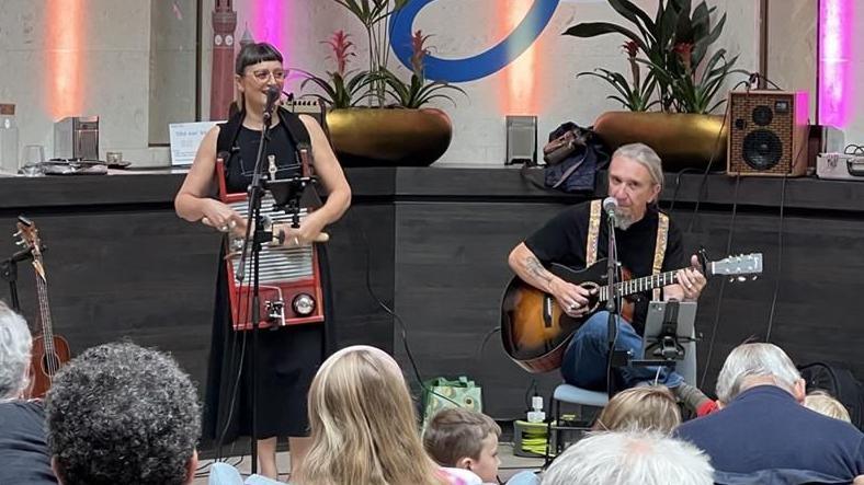 A woman dressed in black and wearing glasses is playing an instrument while singing into a microphone. Next to her a man with long grey hair tired back into a ponytail is sat down while playing the guitar and singing into a microphone as well. In the foreground, the back of the heads of audience members are just visible