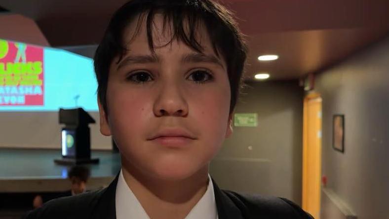 A young boy looking at the camera. He has dark hair that covers his forehead. He has a dark blazer on, with a white shirt and stripped tie. He is in a school hall