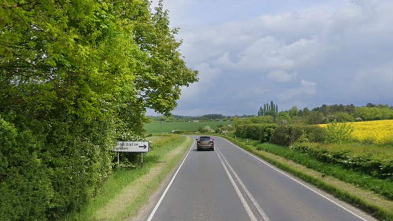 Google image of a road surrounded by greenery and fields. On the left there is a sign for Barnardiston and Huntingdon. There is also a grey driving car on the road away from the camera. 