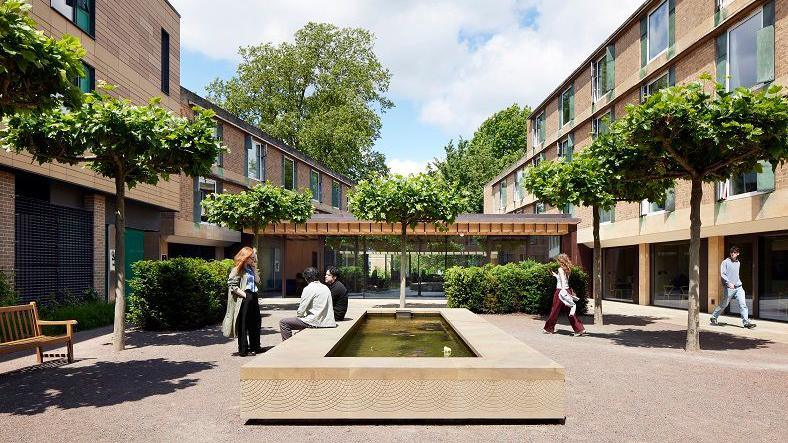 Outdoor area at a modern university building which has four storeys on the right and three to the left. In the centre of a paved area is a rectangular structure with water in the centre on which two people are sitting. There are trees dotted around the area and three people are walking around.