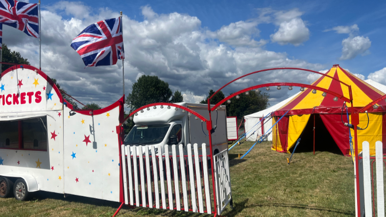 A wide shot of the circus with a white picket fence entrance and a ticket booth painted white with coloured stars on it. The Big Top is in the distance standing in a field with other white tents in the background.