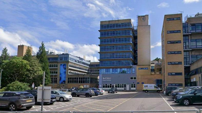 Google Maps image of a large multi-storey university building made up of beige brick with blue window frames and grey cladding with "University of Surrey" written on the front and a carpark in the foreground.