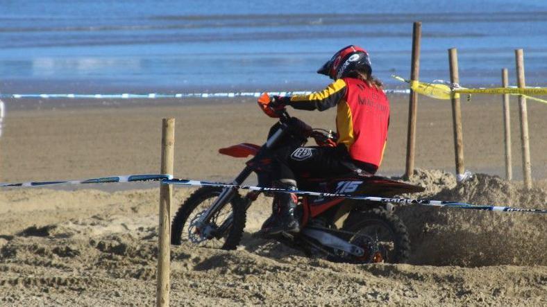 Rear view of a motocross rider trying to turn a corner in deep sand. This rider has black boots and trousers with a red, yellow and black long-sleeved top and a black helmet with red goggles. They are about to make a right-hand turn on the course as sand flicks into the air from their rear tyre.