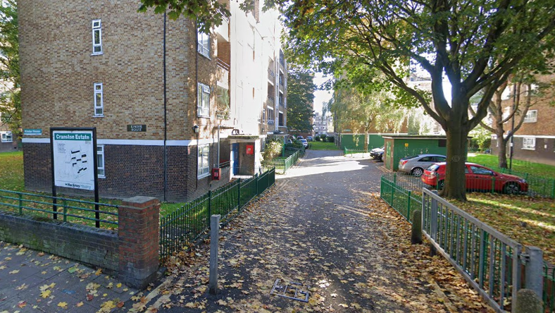 A view of the exterior of one of the buildings at the Cranston Estate in east London. A sign for the estate is visible on the left and cars are parked down a road to the right of the building