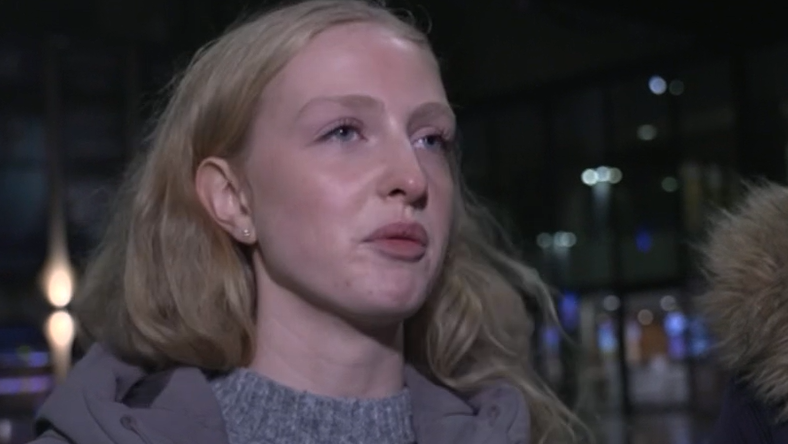 A woman with long blonde hair is being interviewed for the BBC. She is stood near Centenary Square and she wears a thick grey coat and a grey jumper.