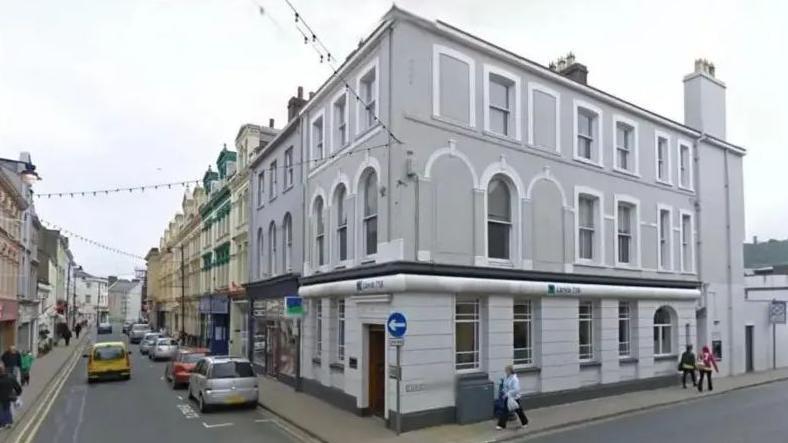 A small town corner, with a grey Victorian building placed on the junction between two streets. The photo shows the street down the left lined with parked cars. 