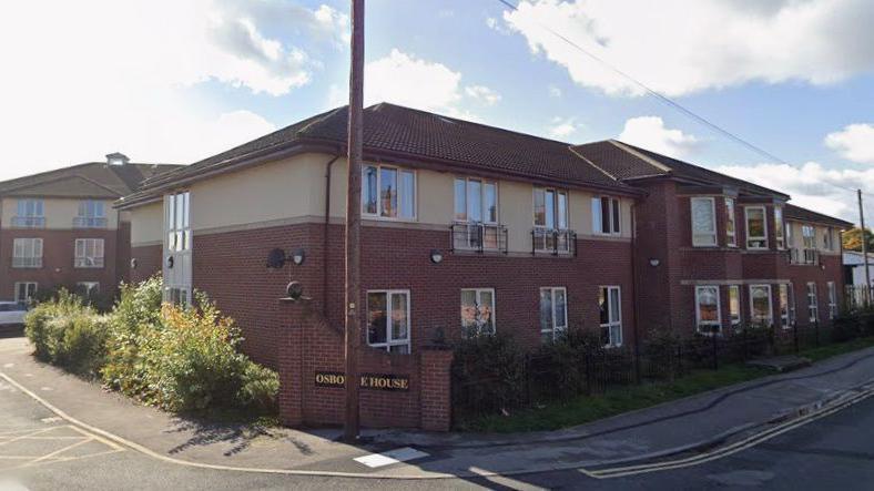 A two-storey brick building with cream-coloured upper walls and a brown tiled roof, identified as Osborne House by a sign near the entrance. The structure has multiple windows, some with small balconies, and is surrounded by low shrubs and greenery.