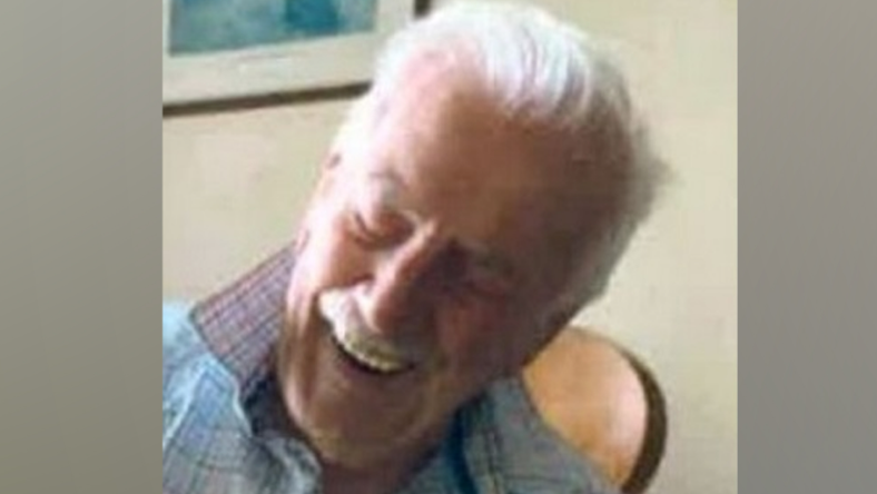 Mr Monk is sitting in a chair with a checked shirt on and is smiling. He has grey hair and a moustache.