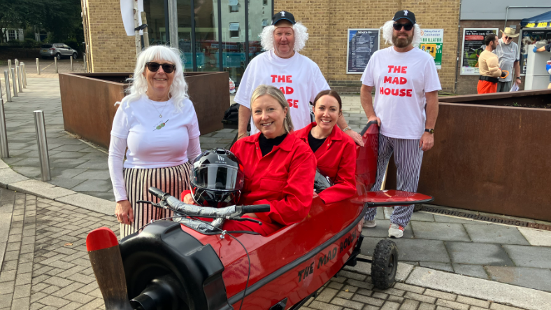 Two women are smiling at the camera and are dressed in red sat in a small kart which is of a red plane. Three people are stood behind them wearing a white top which says THE MAD HOUSE on it. 