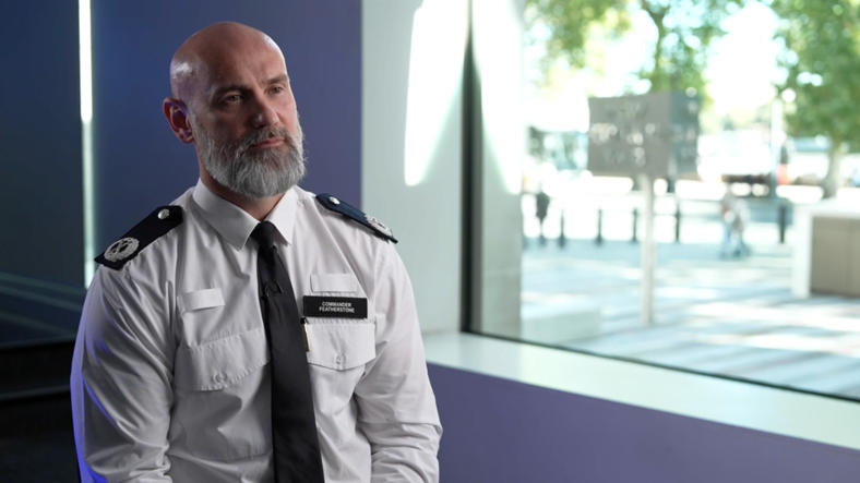 Police officer in uniform sits in front of a window looking off to his right. He is bald with a grey beard