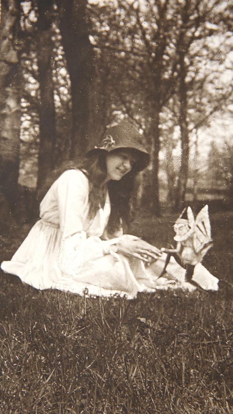 A black and white photo of a young girl with long hair sitting on grass with trees in the background. She is wearing a white dress and a dark hat with a flower on the side and she is smiling while looking at a small mythical creature with wings and a foot in the air.
