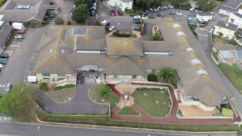 An aerial view of a grey/brown building surrounded by car parks and a main road. There is some greenery in front of the building and there are houses to the right of it.