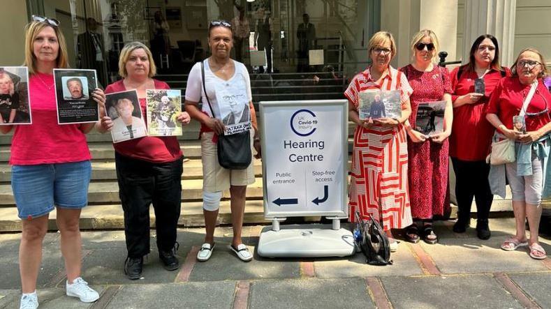 A group of seven people stand outside a large building, with pillars and steps outside that lead to glass doors. They are holding photographs of their relatives who died with Covid during the lockdowns, which began in 2020.