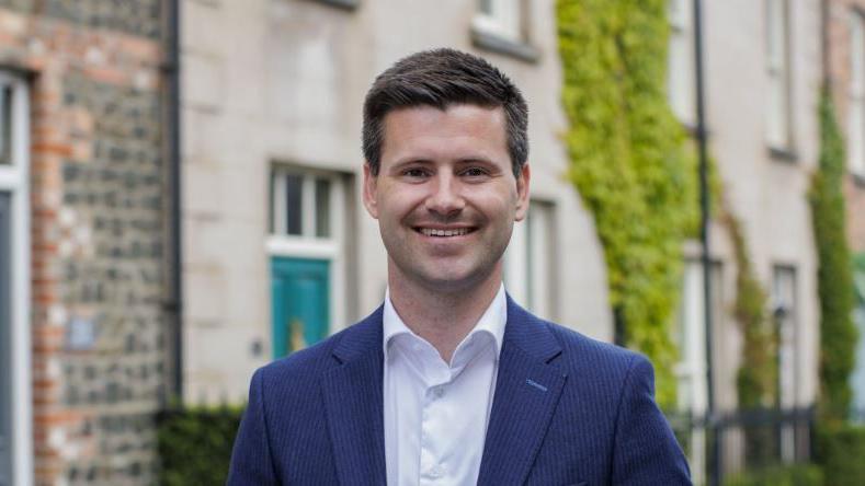 Jonathan Buckley has short black hair and is wearing an open-necked white shirt and blue suit jacket,. A row of terraced houses is in the background