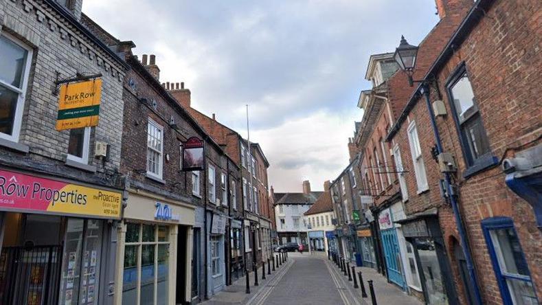 A narrow paved town centre street, complete with shop fronts, pavement bollards and other street furniture.