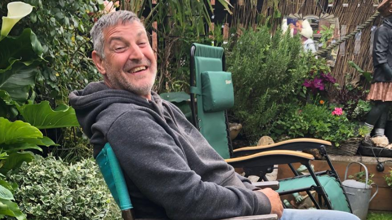 Lee Mainwaring sits on a chair in a garden, surrounded by plants, wearing a grey hoodie and jeans and smiling