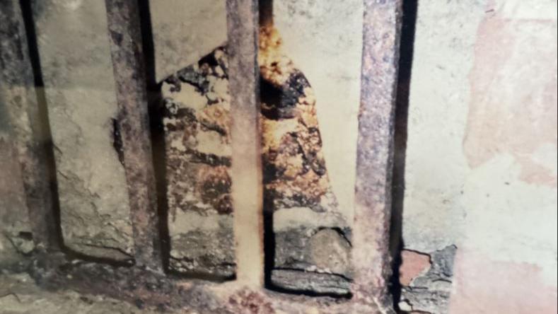 A rusted metal grille with a cracked stone wall behind it