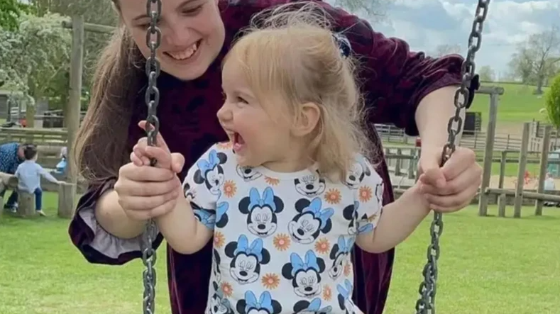 Woman pushing a laughing little girl in a Mickey Mouse dress on park swings