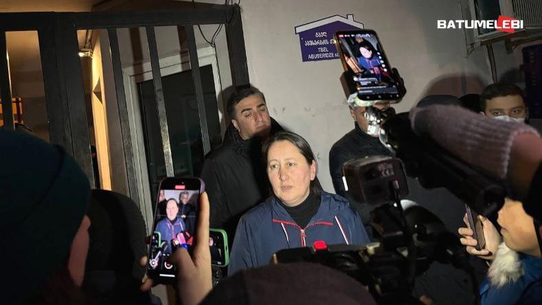 A woman is surrounded by photographers, wearing a blue coat. A mobile phone in the foreground shows her image