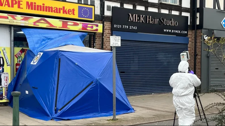 A blue tent covers the crime scene which is outside a vape shop on Station Road. A person in a white overall covering all their body holds an instrument on a tripod in the foreground. The tent is on concrete paving.