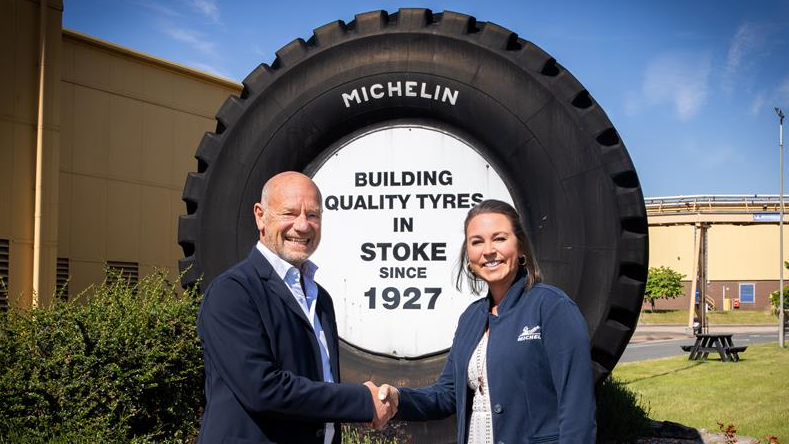 Two people stand in front of a large Michelin tyre, shaking hands. The middle of the large tyre has black text on a white background saying "building quality tyres in Stoke since 1927."