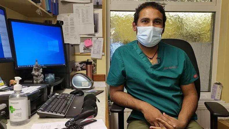 A man with short, black hair and a nurse's green top is sitting in a black office chair wearing a blue mask with his hands in his lap. Next to him is a desk with a computer, keyboard, hand sanitiser and several bits of paper attached to the wall.