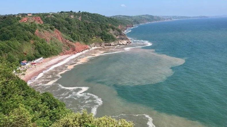 Oddicombe Beach in Torquay - blue sky and sea. Red rockface and green, vegetation covered cliffs