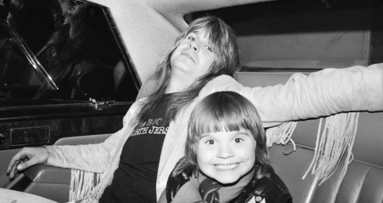 A black and white picture of a man with long hair, a fringed jacket and dark t-shirt with a smiling younger child both in the back of a car.