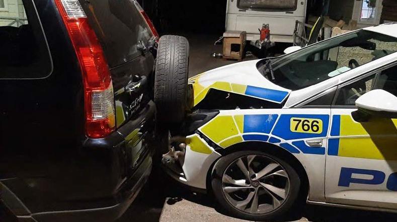 Image shows a black Honda CR-V ramming backwards into a police car. The hood of the car is significantly damaged with a large crack near the wheel.