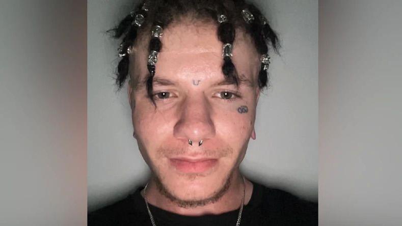 A man taking a photo of himself stood against a white background. He is wearing a black t-shirt, has a tattoo below his left eye, a nose ring and his hair tied in bunches