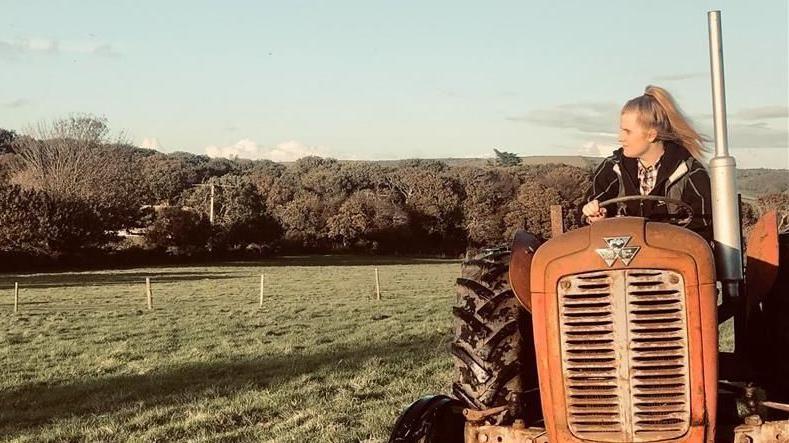 A woman on a farm driving a tractor on a sunny day. She is looking away from the camera.