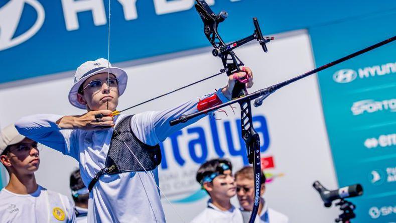 A picture of Baptiste Addis using an archery bow and wearing a white hat and glasses. 