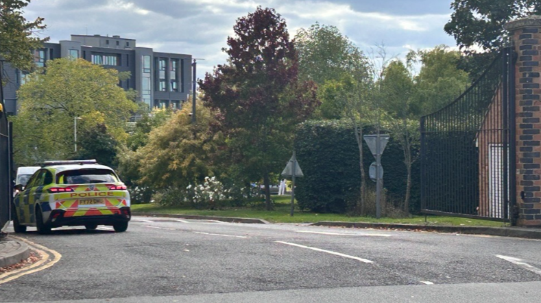 A police car is pulled up at the side of the road, just in front of a large black gates