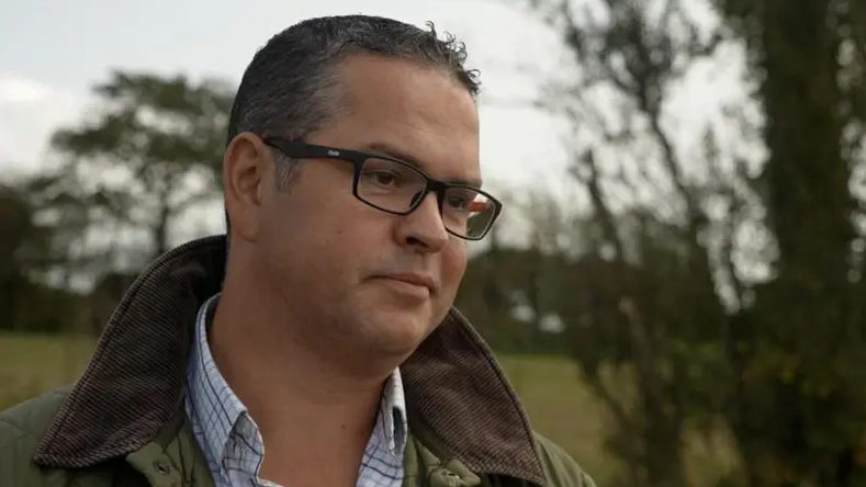 A man with short black hair and rectangle black glasses, wearing a khaki coat with a brown rimmed collar and a blue checkered collared shirt underneath. He is standing outside with trees in the background, which is blurred.