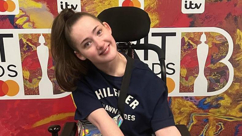Grace Wolstenholme smiling straight into the camera. She is in front of a branded wall with the logo for the Brit Awards 2024.