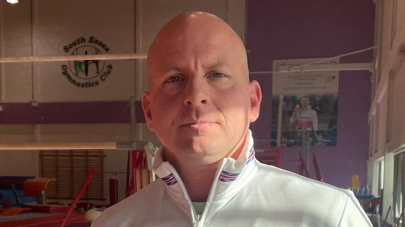 Scott Hann has a shaved head and is wearing a white Team GB jacket with tall collars. He is standing on the floor of a gymnastics club, with a pole behind him.
