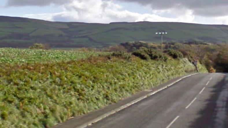 A countryside road with empty overgrown fields and a hill in the background.