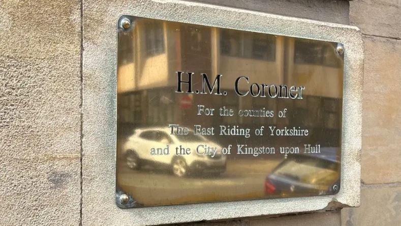 A gold plaque on a white and sandy wall that reads 'H.M. Coroner for the counties of The East Riding of Yorkshire and the City of Kingston upon Hull'.
