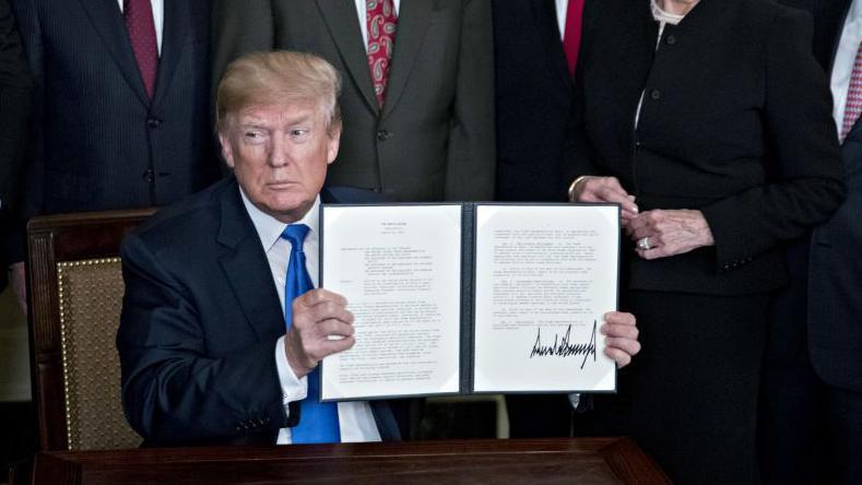 US President Donald Trump holds up a signed presidential memorandum targeting China's economic aggression with a large signature in the Diplomatic Room of the White House in Washington, DC on Thursday, 22 March, 2018. He is wearing a navy suit, white shirt and blue tie. A group of people - whose heads are cut from the photo - stand behind him.