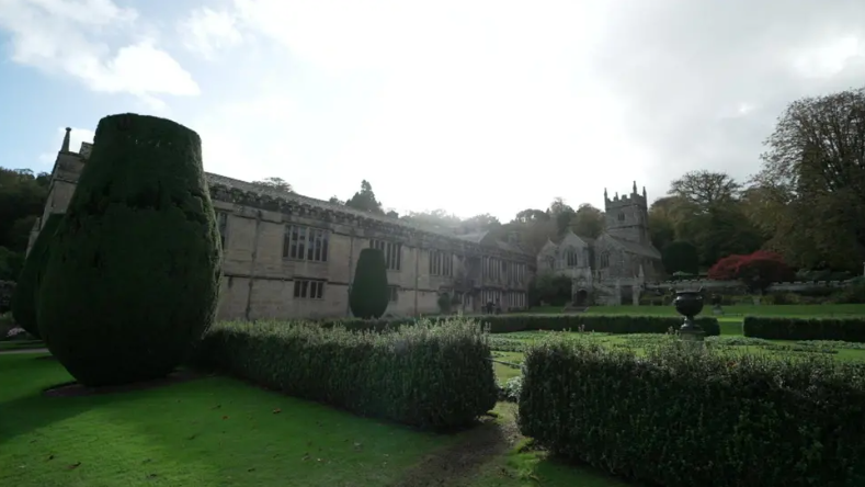 Lanhydrock seen with bushes surrounding the building, there is a castle-type building on the right with turrets, there is a large hedge on the right which has been shaped