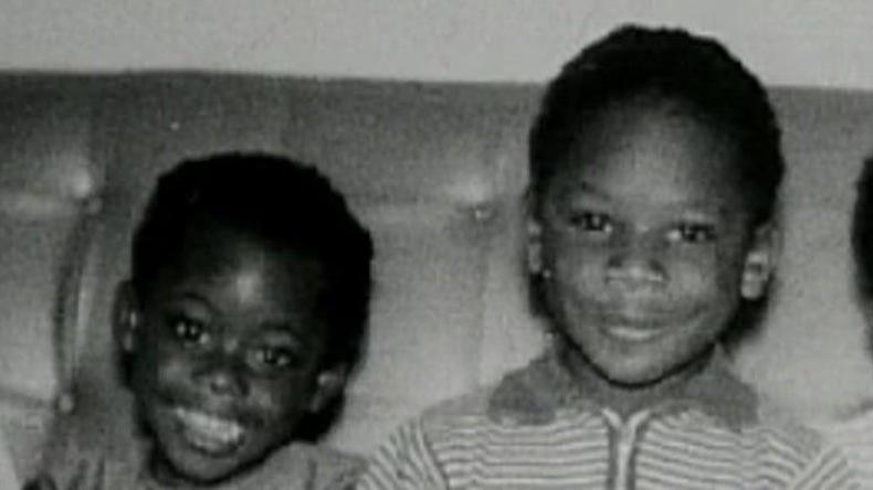 A black and white photo of Janet and Christopher sitting on a leather sofa when they were children. Janet is on the left wearing a capped-sleeve dress and Christopher is on the right wearing a striped t-shirt