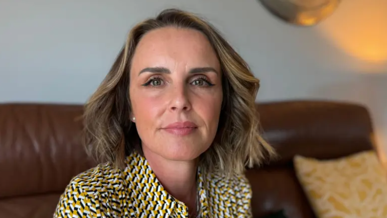 Rachel Williams looks into the camera. She is wearing a yellow, black and white blouse, and is sitting on a brown leather sofa.  